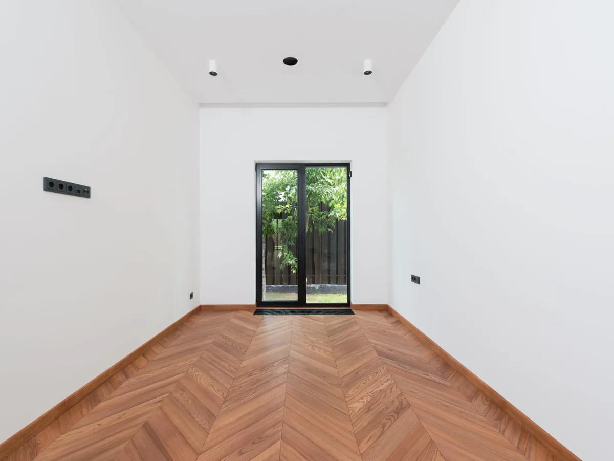 Empty white room with a wood chevron floor whose centre seam runs straight toward a glass patio door
