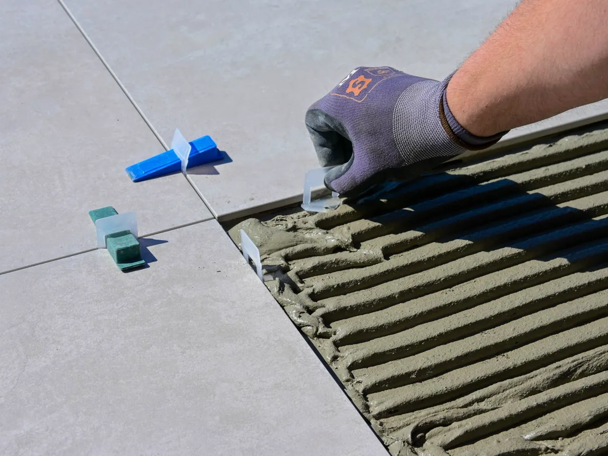 Gloved hand setting a large grey porcelain tile into combed mortar with blue levelling clips